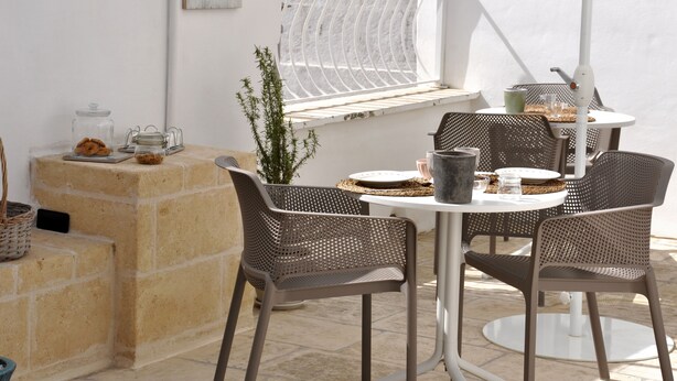 Een terras buiten met een ronde tafel, vier stoelen, een bloempot en een raam met een witte gordijn.