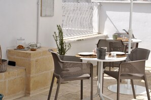 Een terras buiten met een ronde tafel, vier stoelen, een bloempot en een raam met een witte gordijn.