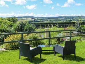 A terrace with two wicker chairs and a table, overlooking a lush garden and a scenic view of rolling hills. - The Wagon House (Launceston)