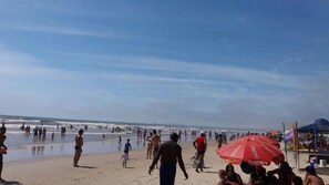 Una playa concurrida donde gente camina y está de pie, y una sombrilla roja con la palabra "TAIPAVA".