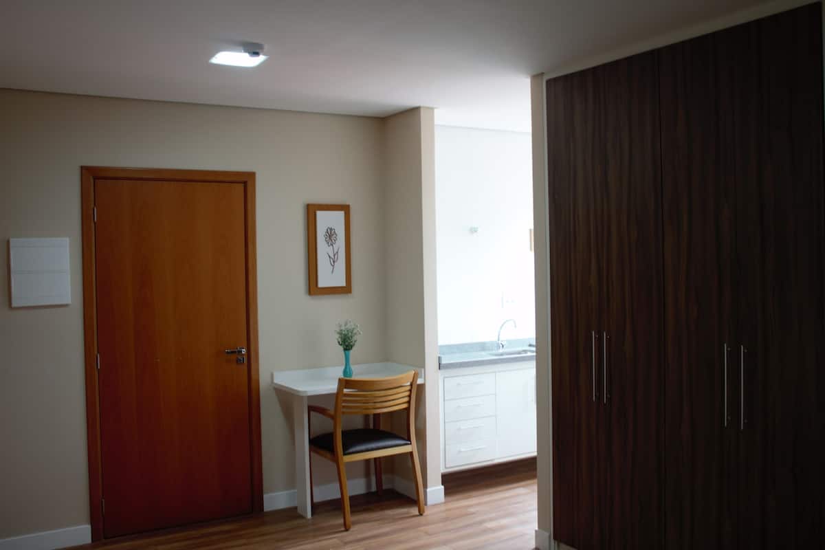 Une pièce avec une porte en bois, une petite table et une chaise, un tableau encadré au mur et une fenêtre donnant sur un paysage.