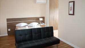 Una habitación de hotel moderna con una cama, una mesita de noche con lámpara, un sofá negro y un aire acondicionado instalado en la pared.