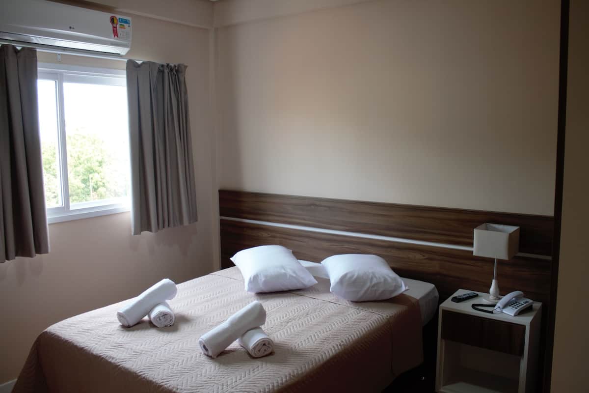 Une chambre d’hôtel comprenant un lit, des oreillers, une table de chevet avec une lampe et un téléphone, ainsi qu’une fenêtre avec des rideaux.