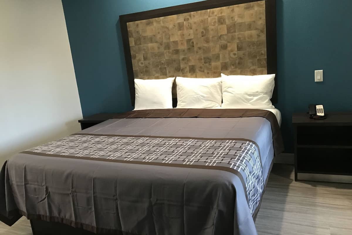 Una habitación de hotel con una cama grande, cabecera con diseño estampado, almohadas blancas, colcha marrón y blanca, y mesita de noche con un teléfono.