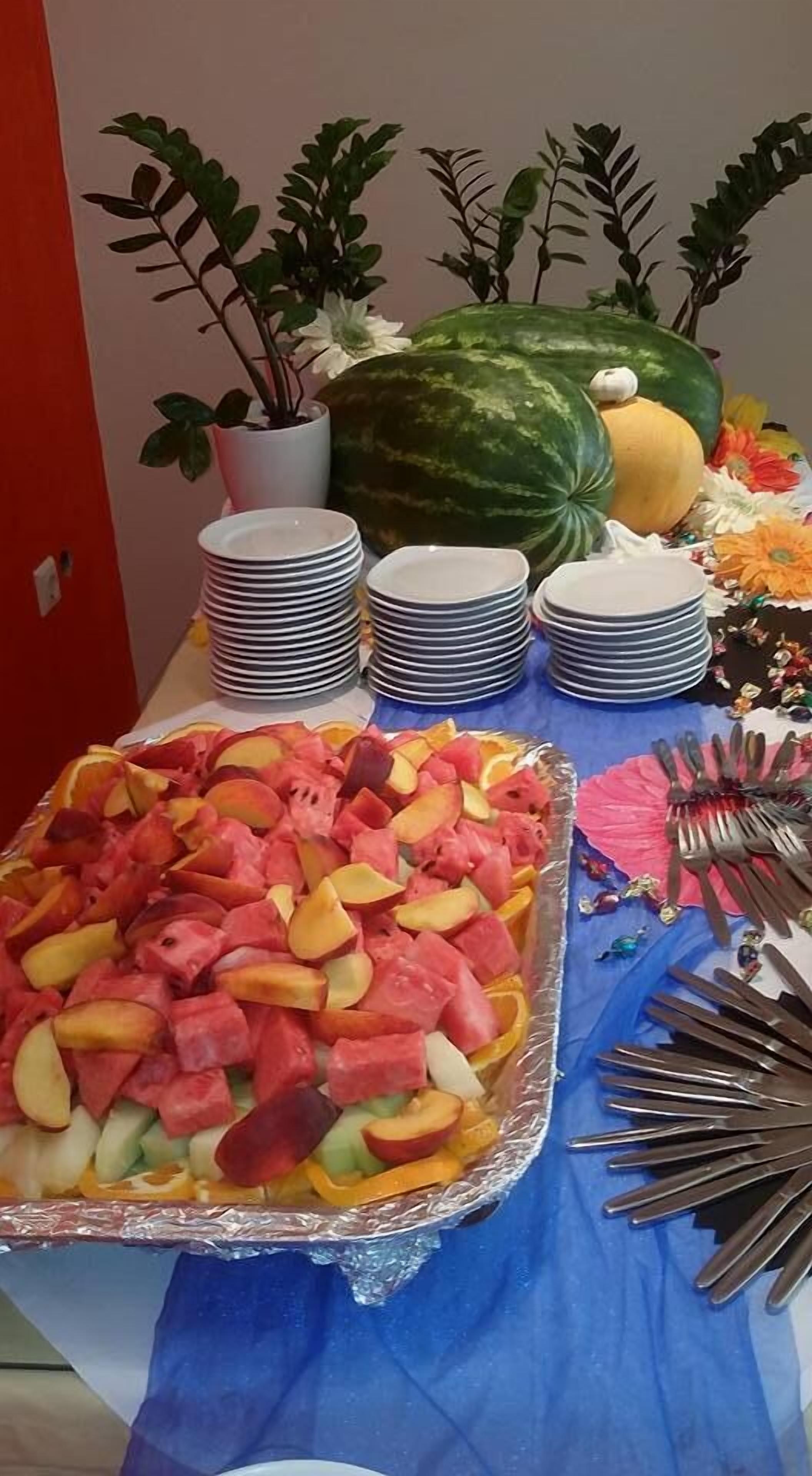Een buffet tafel met diverse soorten fruit en groenten, waaronder watermeloen en meloen, en verschillende potplanten.