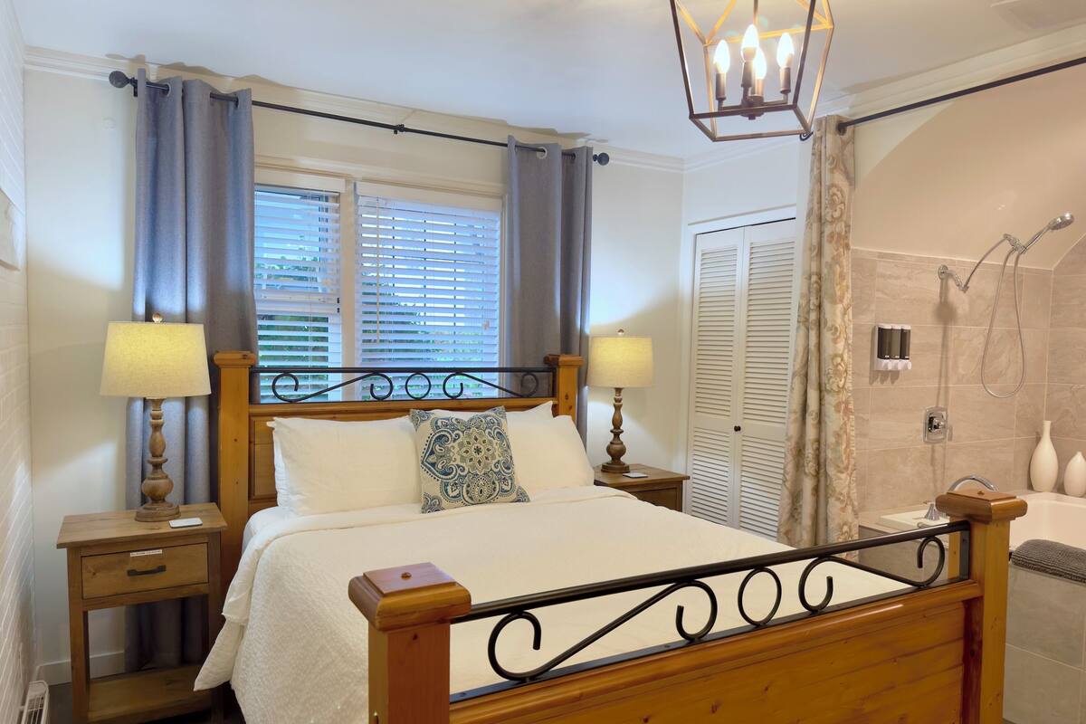 A neatly arranged bedroom with a wooden bed, bedside lamps, a window with curtains, and a shower area.