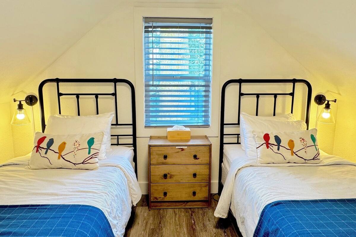 A small, cozy room with two single beds, a wooden nightstand, and a window with blinds.