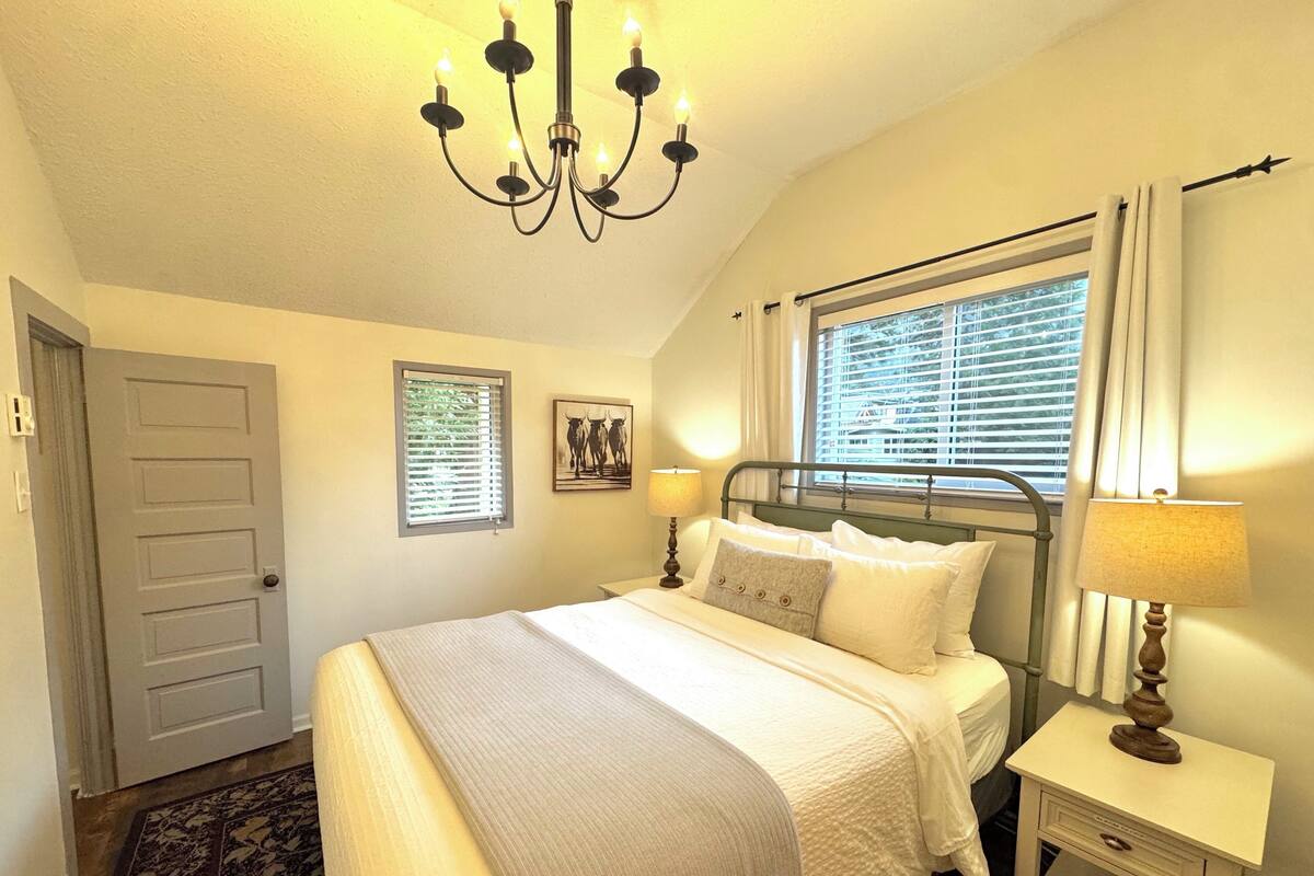 A bedroom with a bed, bedside lamps, a chandelier, a window with blinds, a door, and a small bedside table.