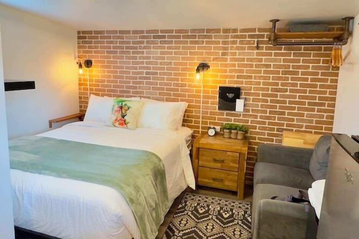 A hotel room with a brick-patterned wall, a bed with a green blanket, a wooden nightstand with a clock, a gray sofa, and a patterned rug.