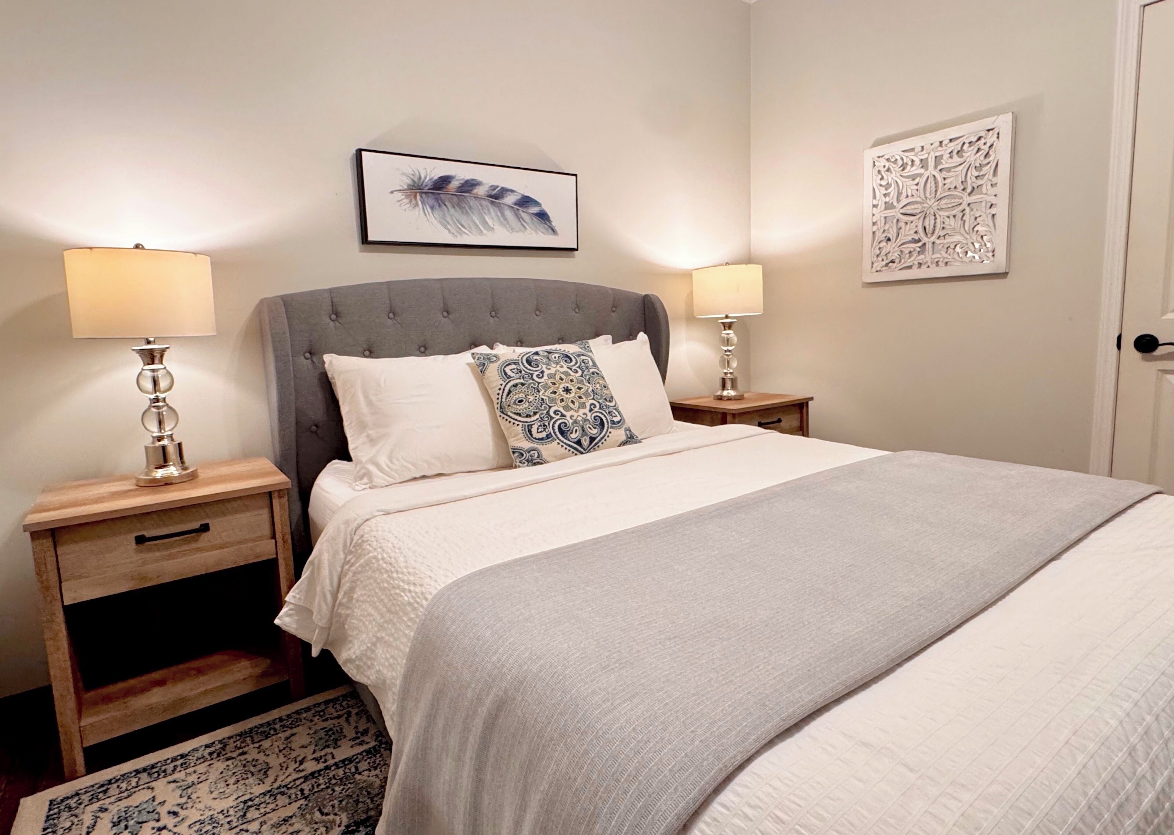 Un lit bien fait, recouvert d’une couverture grise, deux tables de chevet avec des lampes, un coussin à motifs et un élément décoratif fixé au mur.