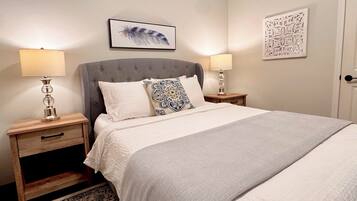Un lit bien fait, recouvert d’une couverture grise, deux tables de chevet avec des lampes, un coussin à motifs et un élément décoratif fixé au mur.