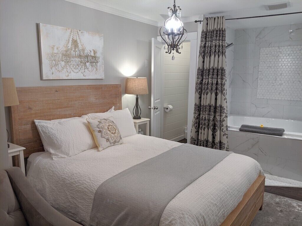 Un dormitorio con una cama grande, cabecera de madera, araña, mesita de noche con lámpara, y un baño con bañera y cortina de ducha.