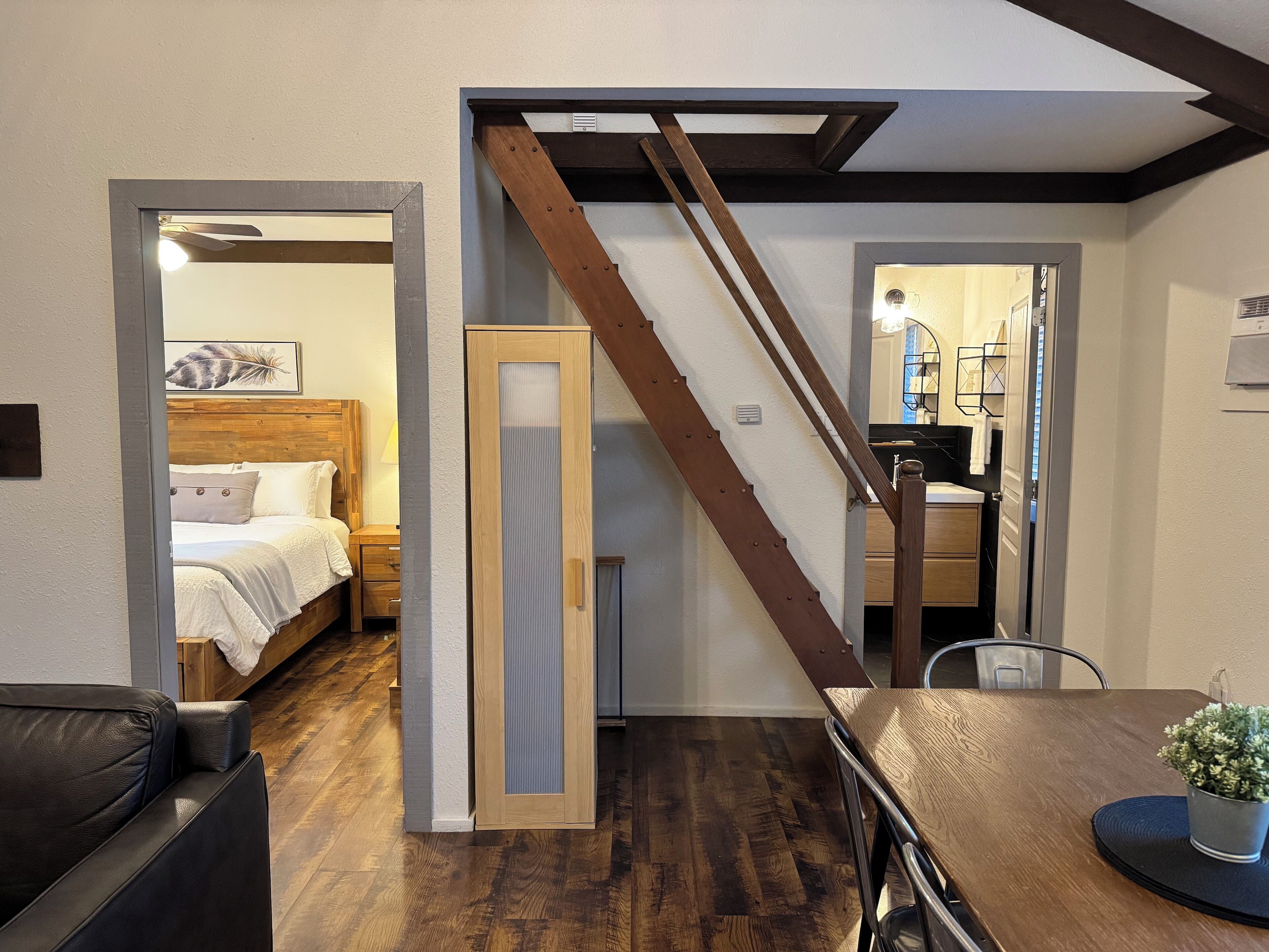 Un dormitorio moderno con cabecera de madera, cama con sábanas blancas y una mesita de noche. Una escalera con barandilla de madera conduce a un baño con lavabo y espejo.