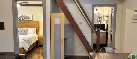 Un dormitorio moderno con cabecera de madera, cama con sábanas blancas y una mesita de noche. Una escalera con barandilla de madera conduce a un baño con lavabo y espejo.