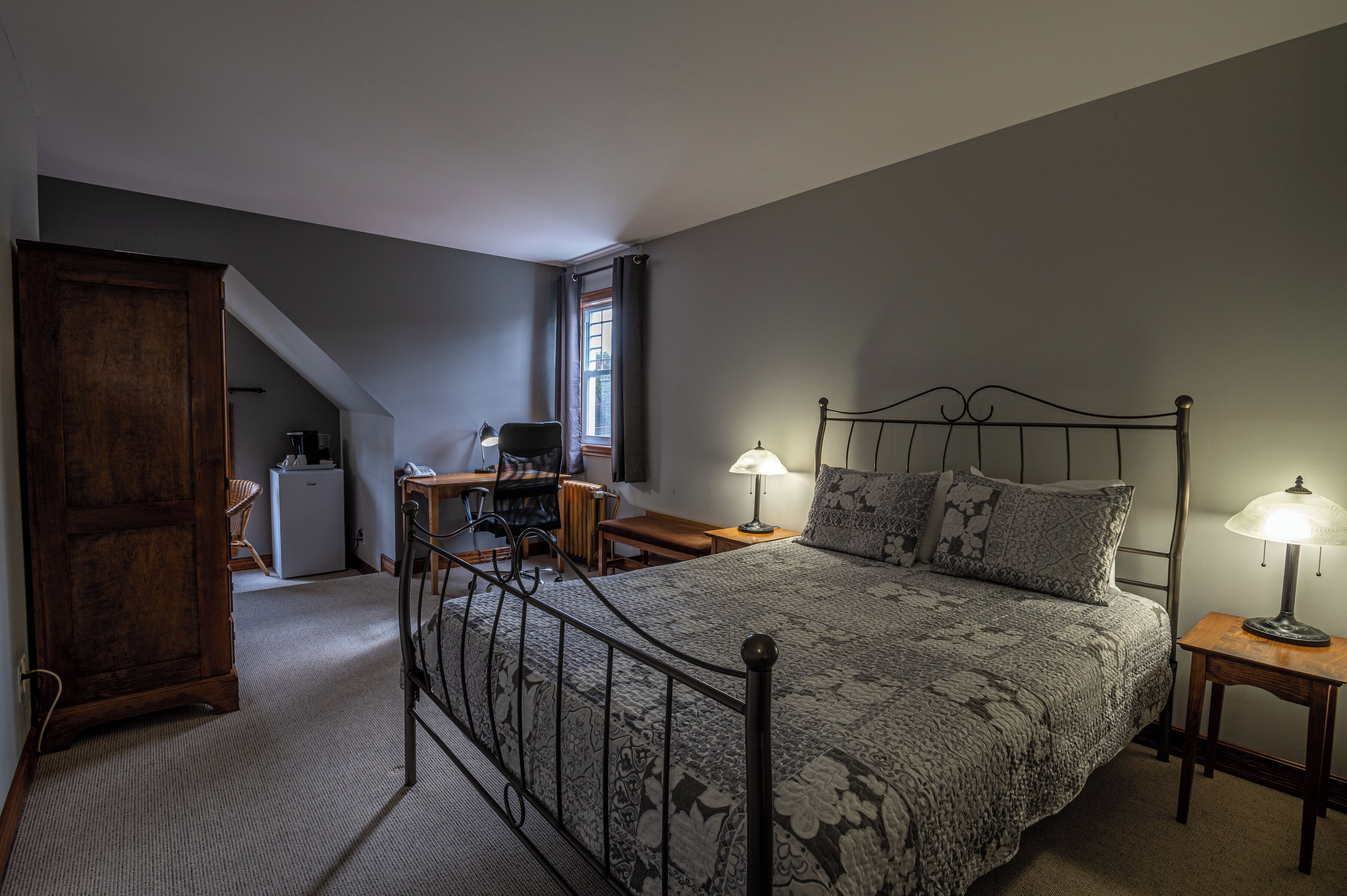 Une chambre à coucher comprenant un lit, deux lampes de chevet, un bureau avec une chaise, un petit réfrigérateur et une fenêtre avec des rideaux.