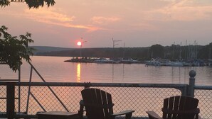 Zonsondergang vanaf een steiger met stoelen en een tafel, uitkijkend over een rustig wateroppervlak met boten en een verre kustlijn.