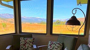 Una zona de estar acogedora con sofá y dos sillones, ventanas amplias con vistas a un campo y montañas, y una mesa.