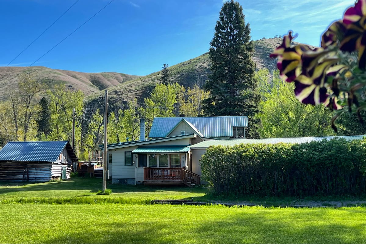 A house with a green roof, surrounded by a well-maintained lawn and a forested hillside.