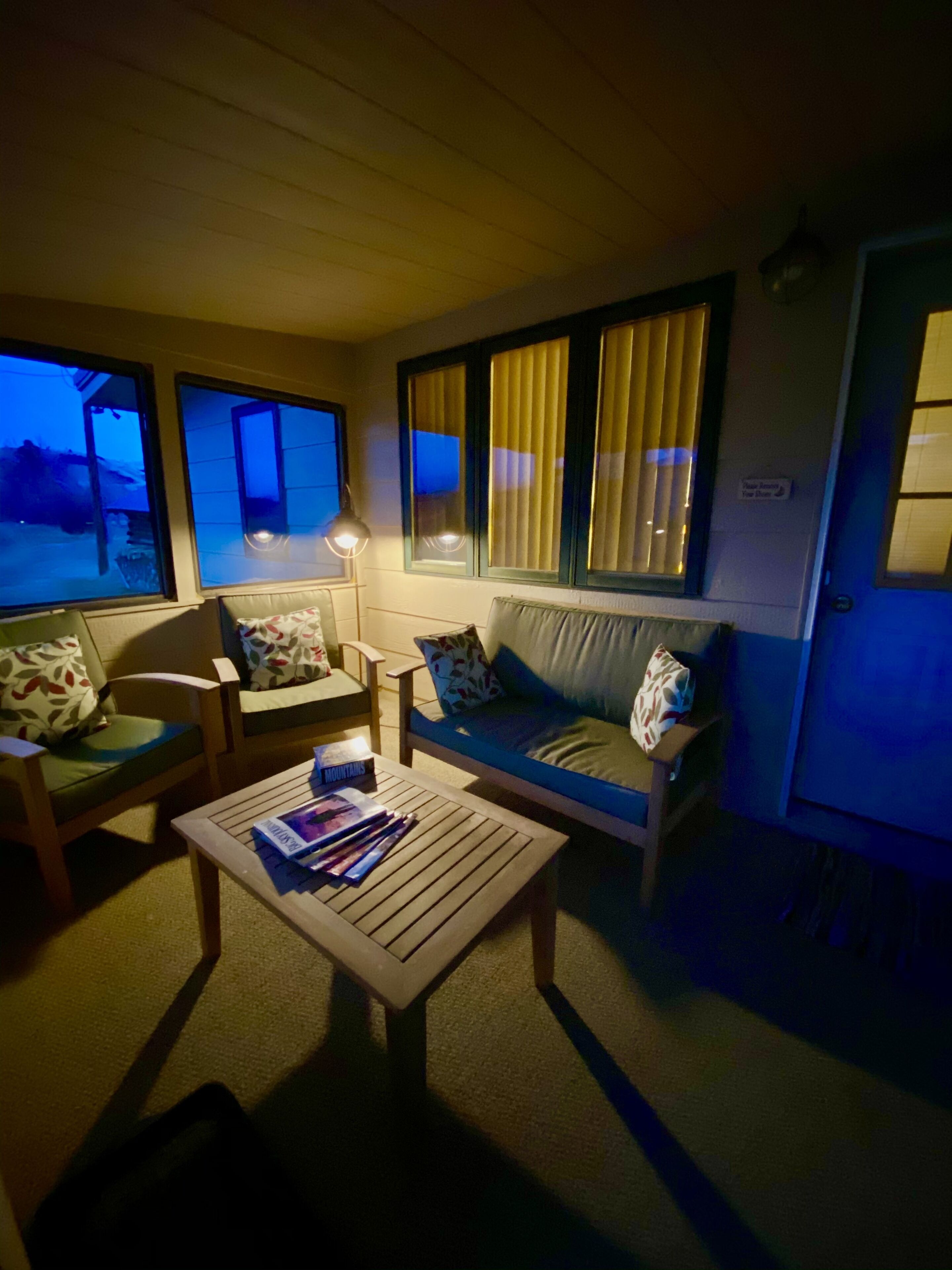 A cozy living area with a sofa, chairs, and a coffee table. There are windows with blinds, a lamp, and a sign on the wall.