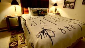 Un dormitorio con una cama con estampado de anzuelo de pesca, una mesita de noche con lámpara, un cuadro colgado en la pared y una ventana con vistas al cielo.