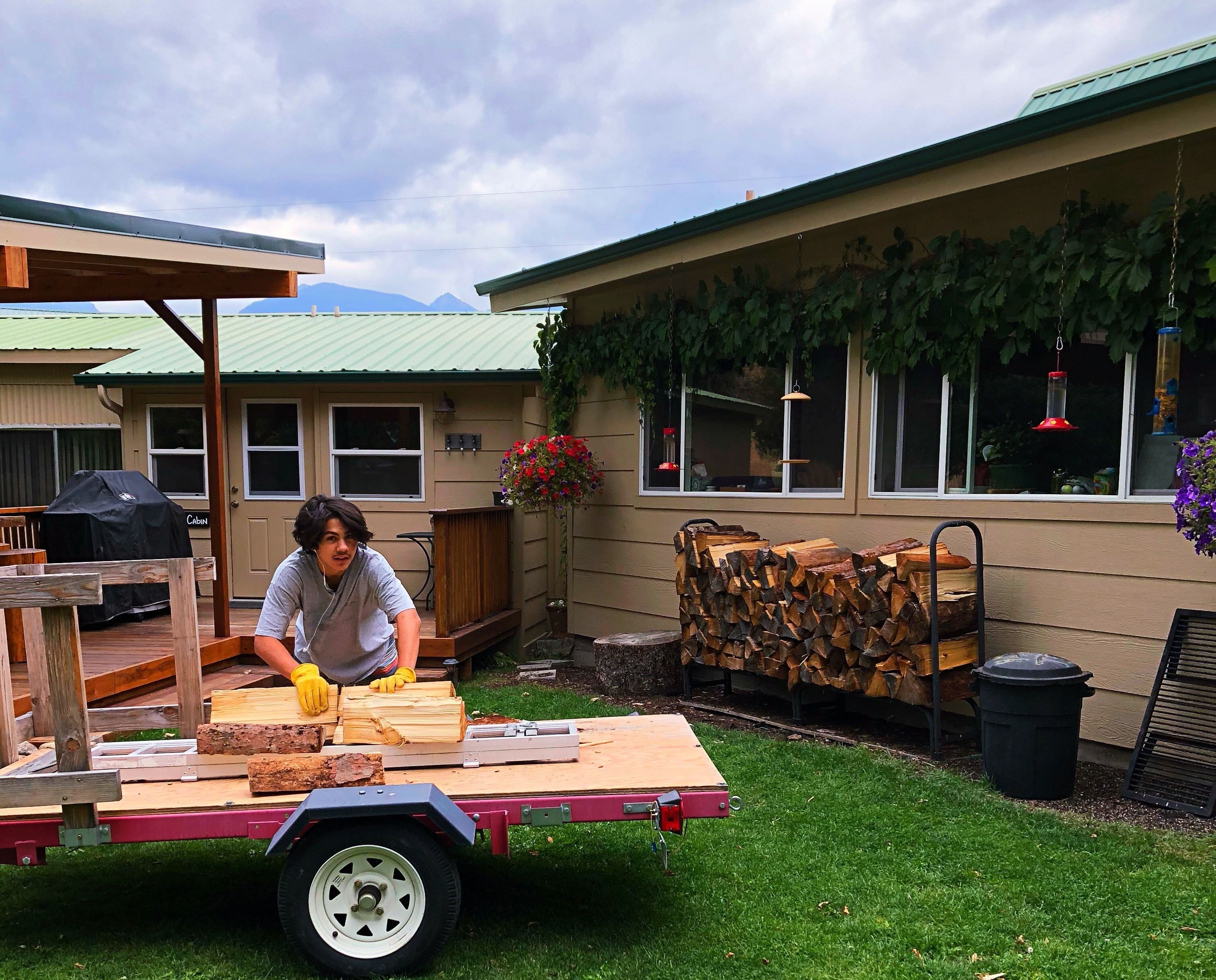 A person working with wood on a trailer in a backyard with a house, a covered grill, and a hanging planter.
