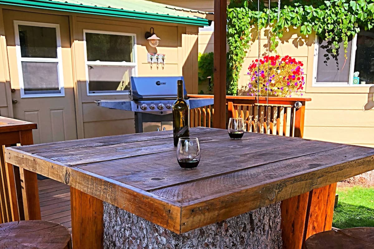 An outdoor wooden table with a bottle and glasses, surrounded by wooden chairs, under a pergola.