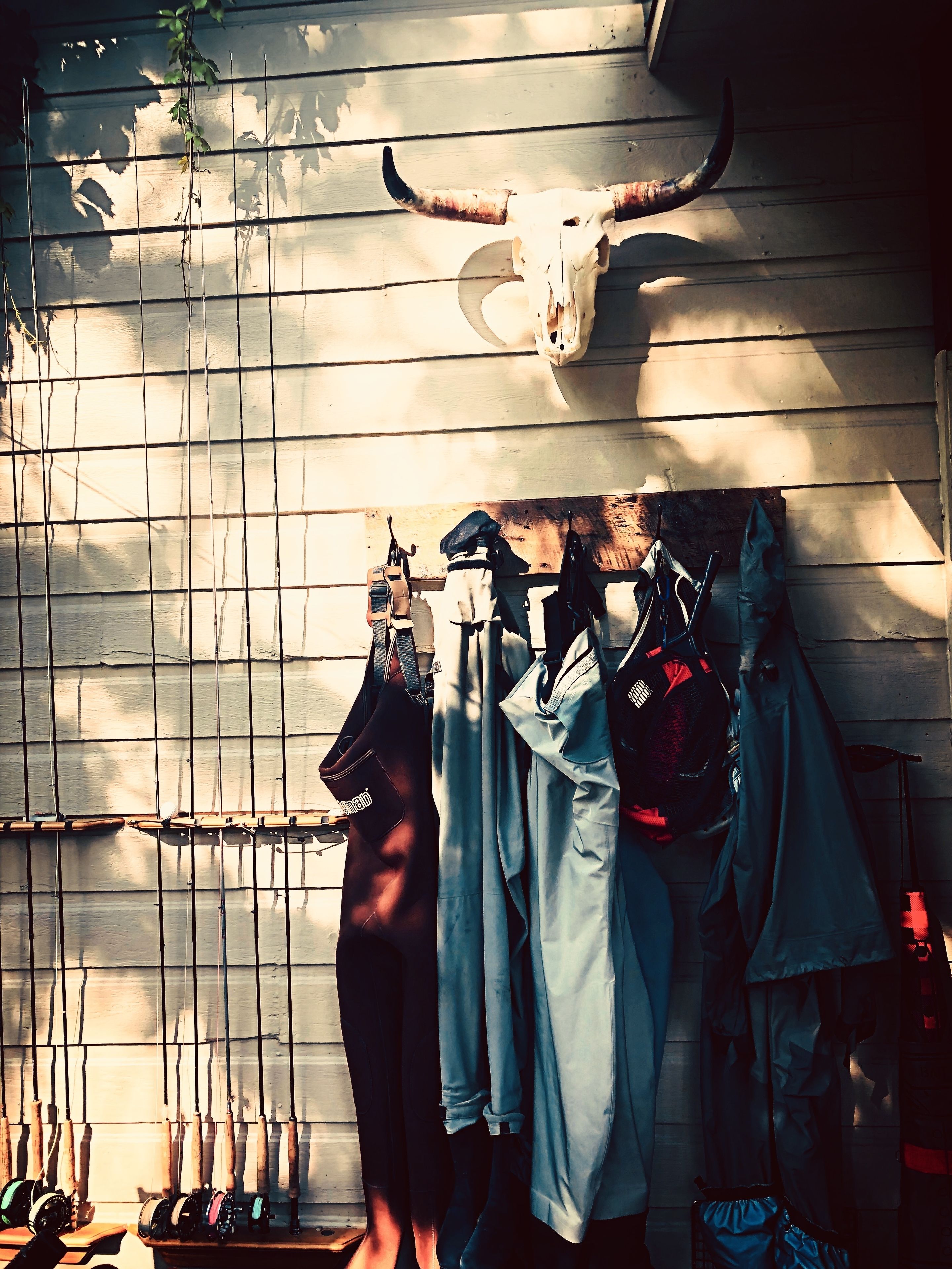A rustic interior with a wooden wall, a mounted cow skull, and various items like fishing rods and bags hanging on a rack.