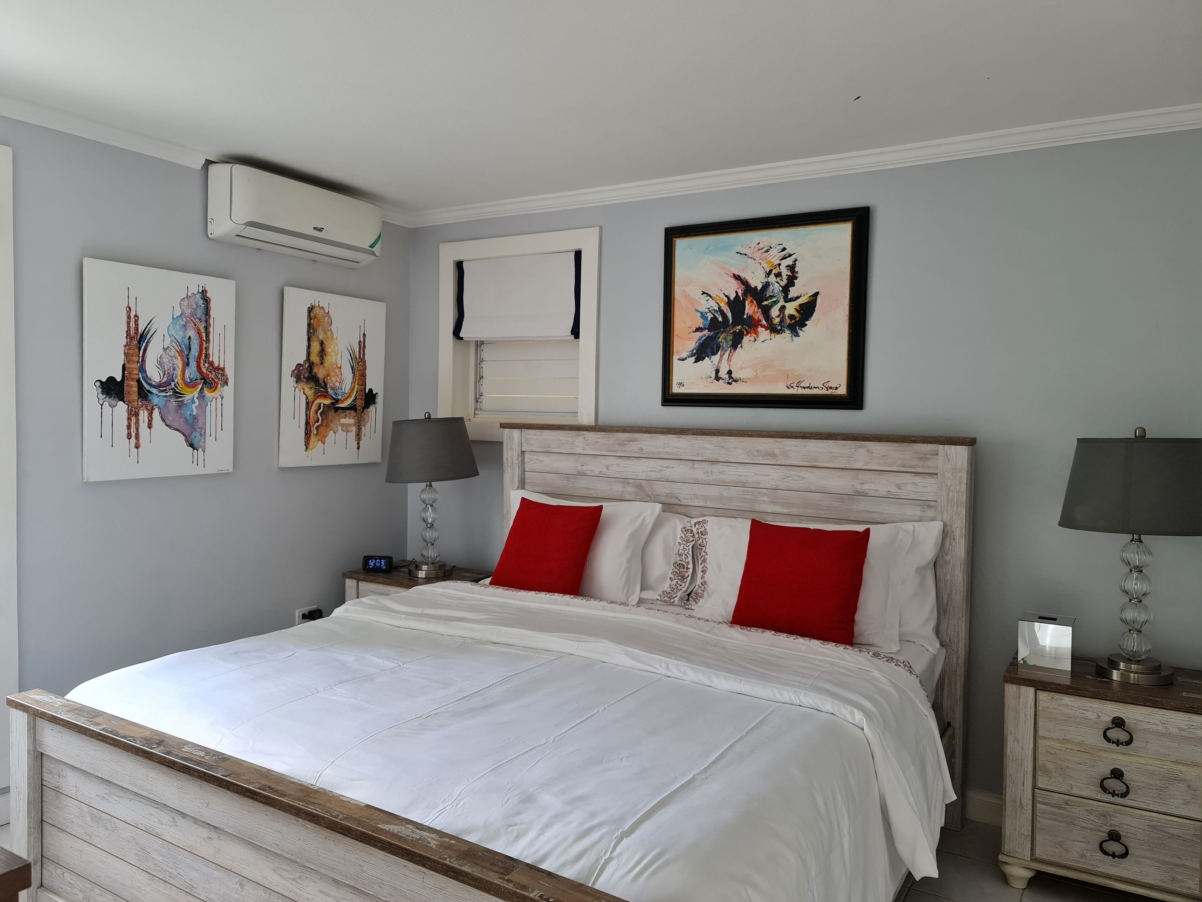 Un dormitorio con estructura de cama de madera, ropa de cama blanca, almohadas rojas, una mesita de noche con lámpara y obras de arte en las paredes.
