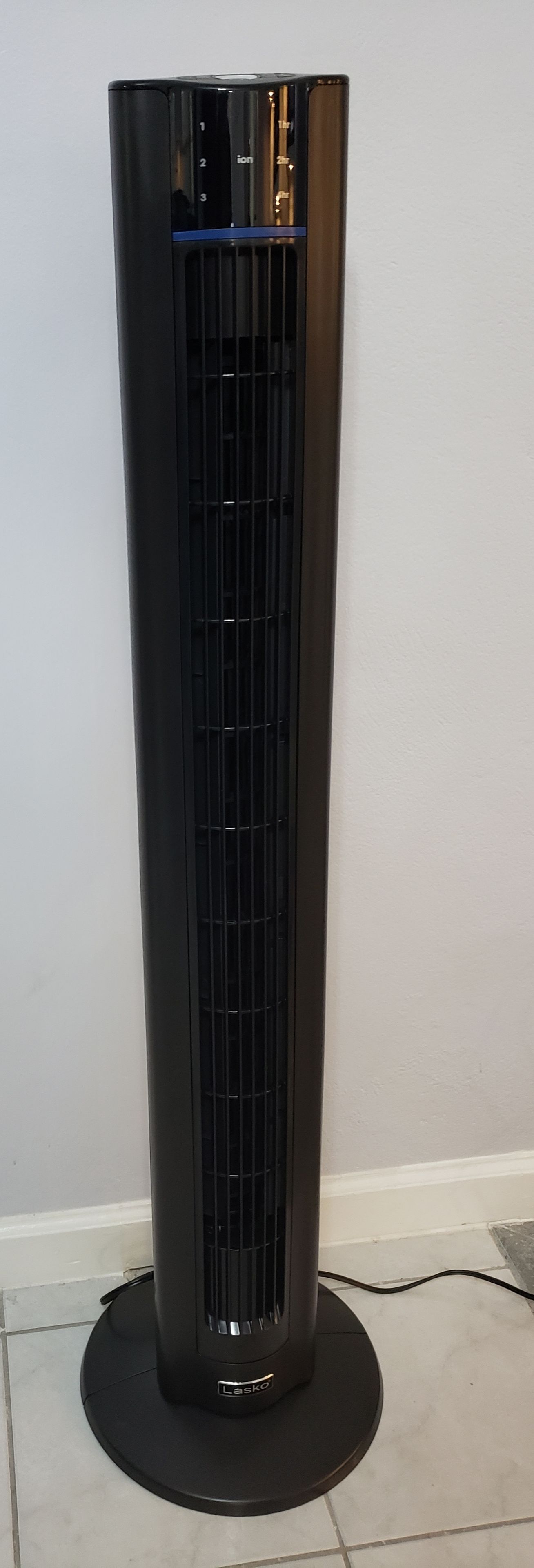 Un grand radiateur portatif noir, doté d’un panneau de commande sur le dessus, est posé sur un sol carrelé, adossé à un mur uni.