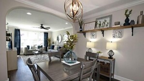Un coin repas avec une table et des chaises en bois, un lustre et un vase avec des plantes. On aperçoit une cuisine à l’arrière-plan, avec un ventilateur de plafond.