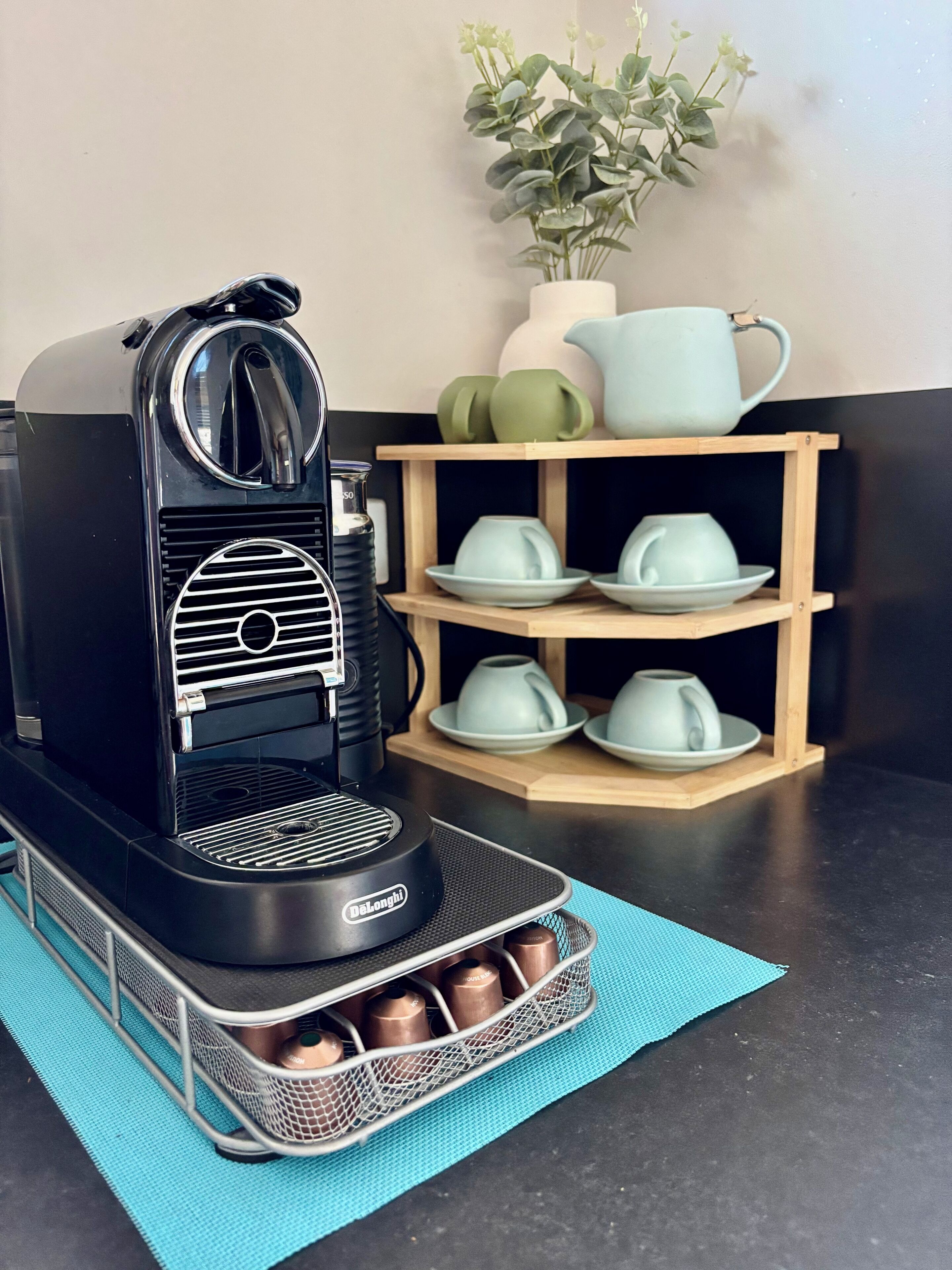 A black coffee machine with a mesh basket for coffee pods, a wooden shelf with cups, and a plant in the background.