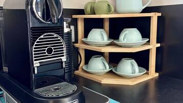 A black coffee machine with a mesh basket for coffee pods, a wooden shelf with cups, and a plant in the background.