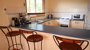 A kitchen with a blue countertop, a coffee maker, a kettle, a microwave, and a stove. There are three wooden chairs with metal frames. - Pegasus Retreat - in the coastal village of Venus Bay (Venus Bay)