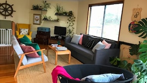 A living room with a sofa, armchair, coffee table, and a baby stroller. There are plants on shelves, a TV, and a window with blinds. - Pegasus Retreat - in the coastal village of Venus Bay (Venus Bay)