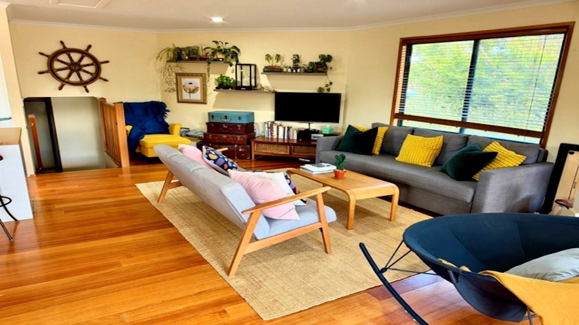 A living room with a sofa, armchair, coffee table, wooden flooring, and a large window with blinds.