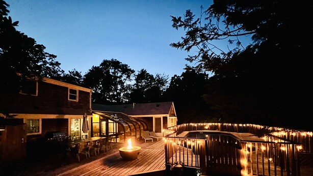 A wooden deck with a fire pit, outdoor seating, and a pool area at dusk. - Private Heated Pool • 5BR/3BA • Near Beaches and Nickerson Park (Brewster)