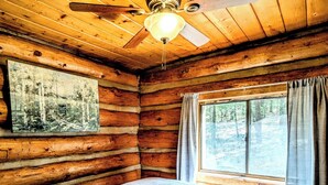 Ein Schlafzimmer in einer Blockhütte mit Bett, Deckenventilator, einem Fenster mit Vorhängen und einem Bild an der Wand.