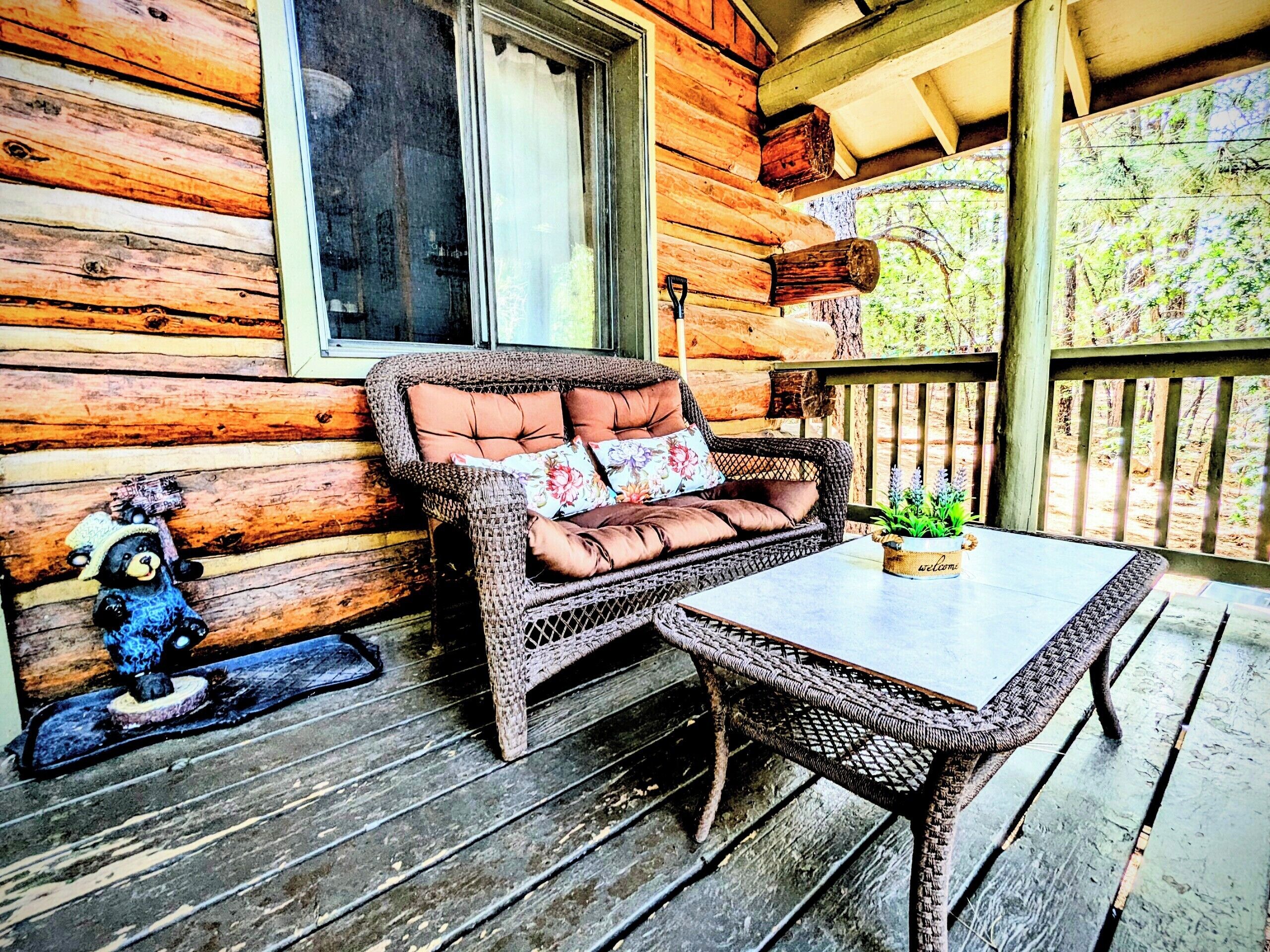 Ein Korbsessel mit floralen Kissen auf einer Holzterrasse, ein kleiner Tisch mit einer Pflanze und eine Bärenfigur.
