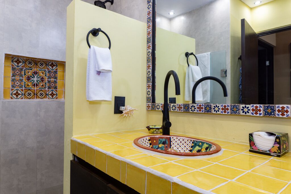 Een badkamer met een geel betegeld wastafelmeubel, een zwarte kraan, een spiegel met een decoratieve tegelrand en een klein plankje met een pak tissues.