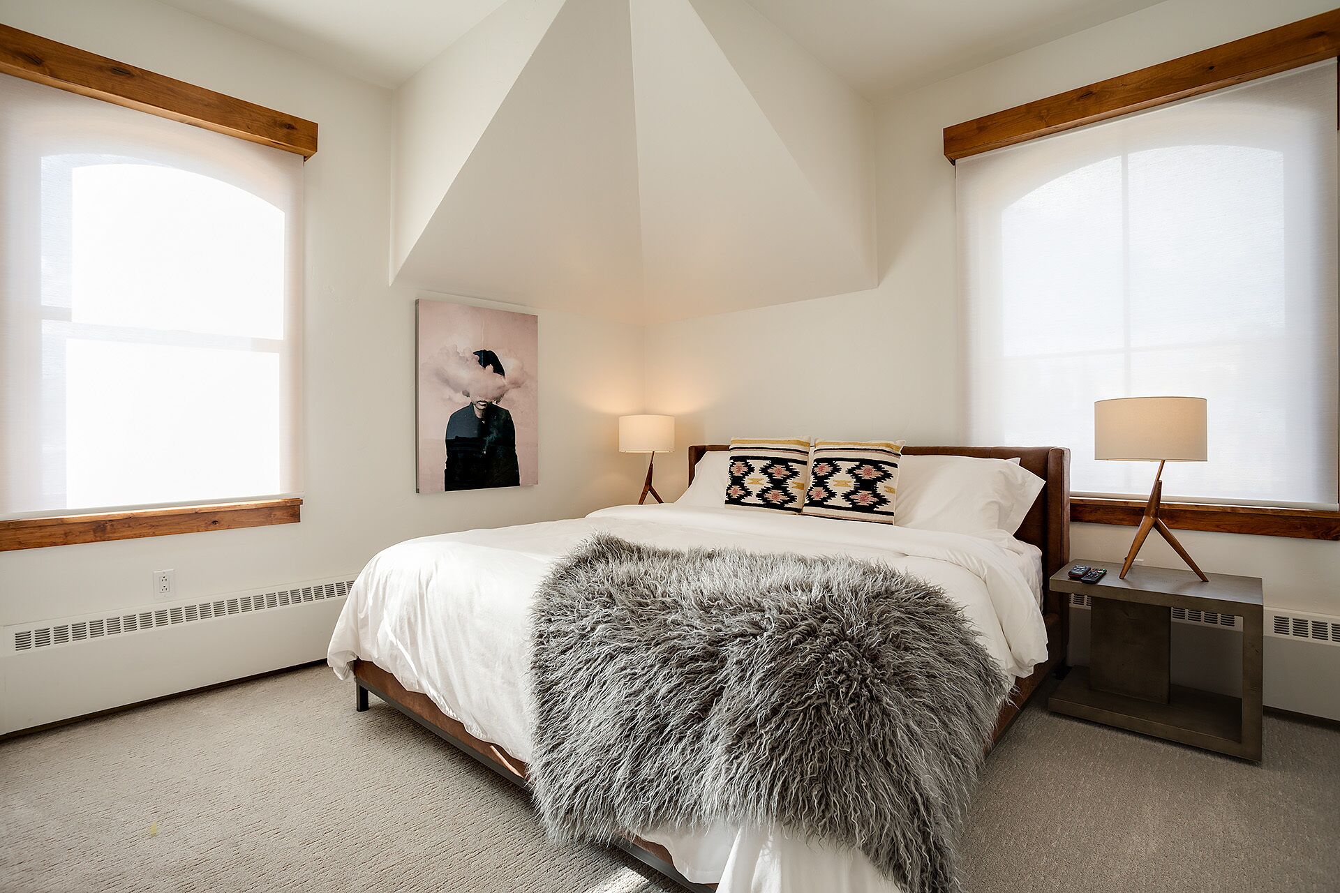 A bedroom with a bed, two bedside lamps, a large window, and a wall-mounted artwork.