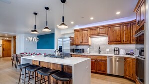 Una cocina moderna con armarios de madera, electrodomésticos de acero inoxidable, una isla central con taburetes altos y lámparas colgantes.