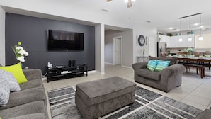 A modern living room with a flat-screen TV, grey sofas, a coffee table, and a dining area with a table and chairs.