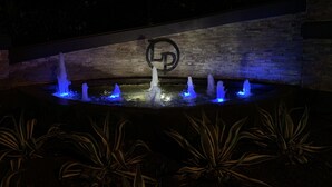 A fountain with water jets illuminated by blue lights, set against a stone wall with a logo.