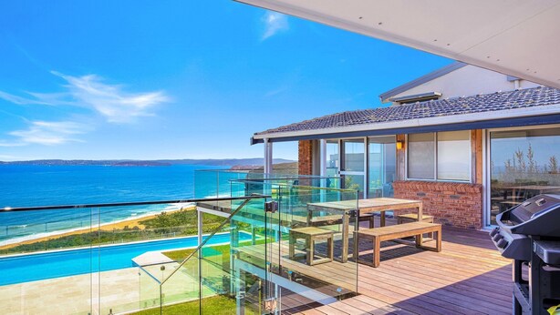 En balkon med gelænder af glas, hvorfra man kan se en pool og havet.