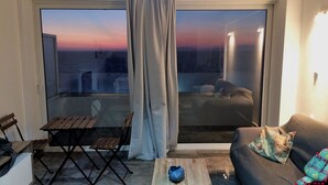 Un salon moderne doté d’une grande porte-fenêtre coulissante donnant sur un balcon. À l’intérieur, on trouve une table basse en bois, un canapé agrémenté d’un coussin décoratif à motifs, et une petite table avec une chaise.
