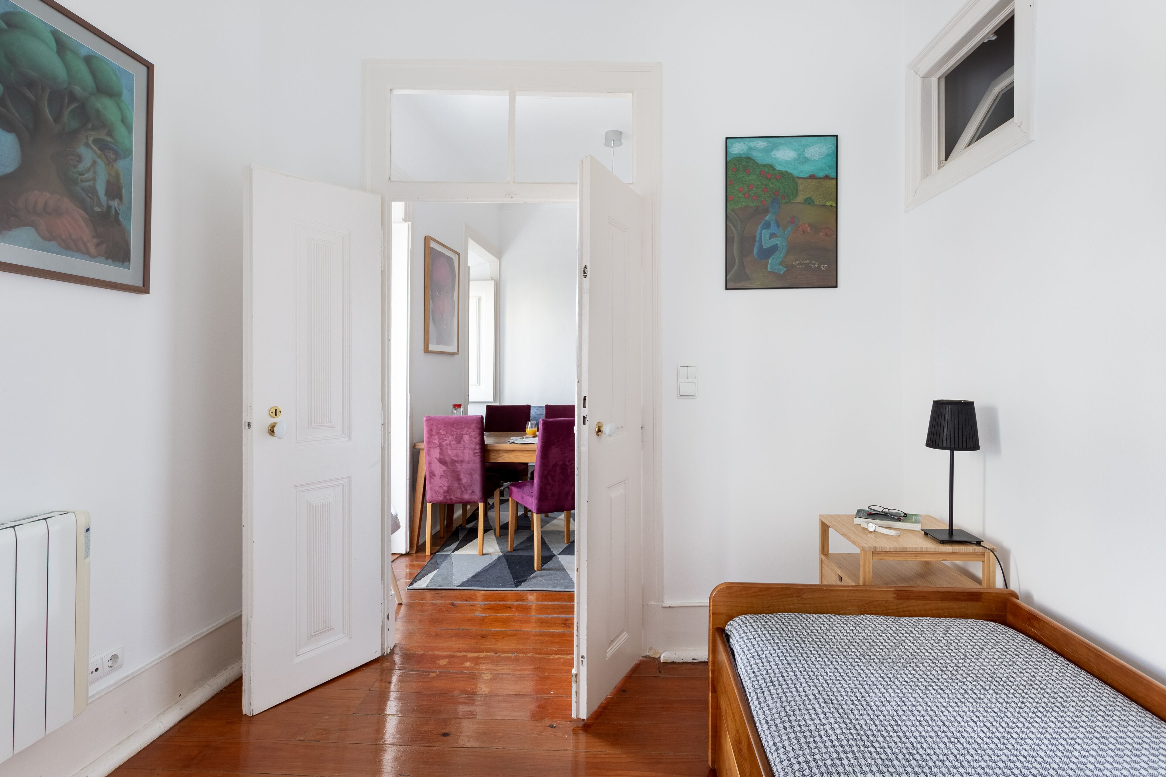 Une pièce avec un lit en bois, une petite table avec une lampe, une chaise et un bureau avec une chaise violette. Une porte donne sur une autre pièce qui contient une table et des chaises.