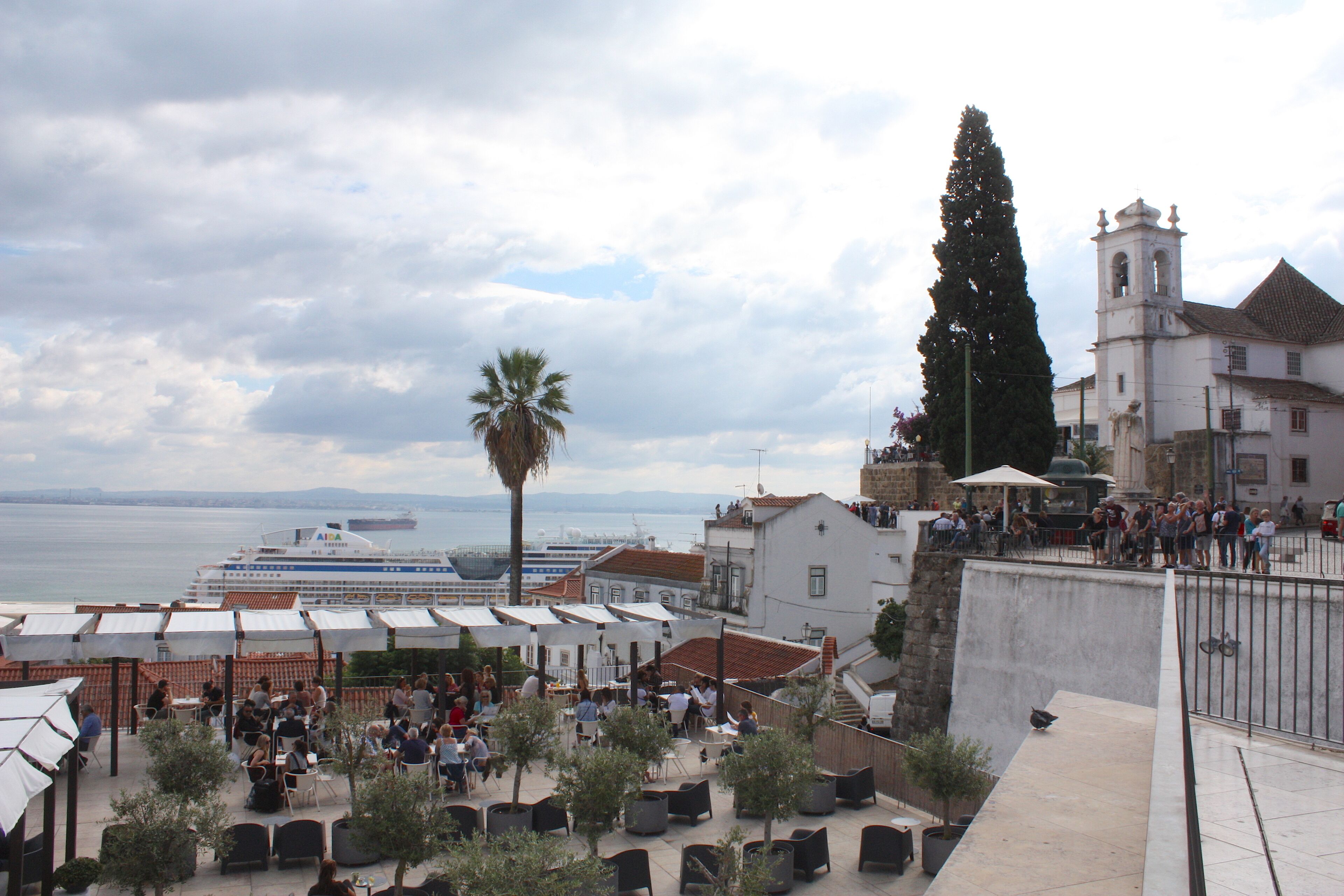 A coastal town with a prominent church, a large ship in the background, and outdoor seating areas.