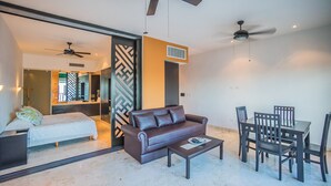 A modern living room with a brown leather sofa, a coffee table, and a dining area with chairs and a table. There is a ceiling fan, a bed with a white bedspread, and a large mirror. - Great 1 Bedroom Studio at Las Brisas by BRIC Vacation Rentals in Playa del Carmen (Playa del Carmen)
