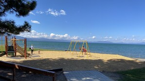 Zona de jocs amb tobogan i cadira de balanceig, una cadira i una àrea pavimentada que condueix a una platja amb una vista clara del mar i muntanyes al fons.
