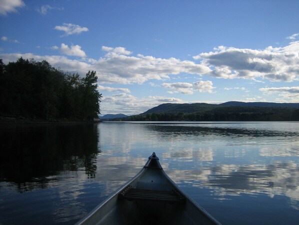 A calm lake with a canoe in the foreground, surrounded by forested hills under a partly cloudy sky.